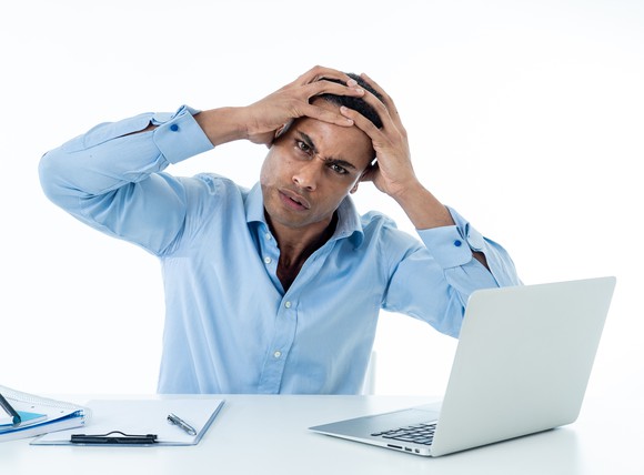 A man with a pained expression holds his hands to his head in front of a laptop.
