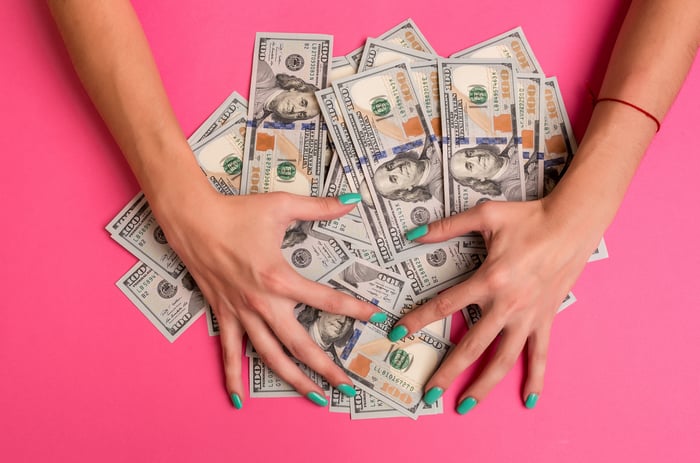 Woman with green-painted nails placing her hands over a pile of hundred-dollar bills against a pink background