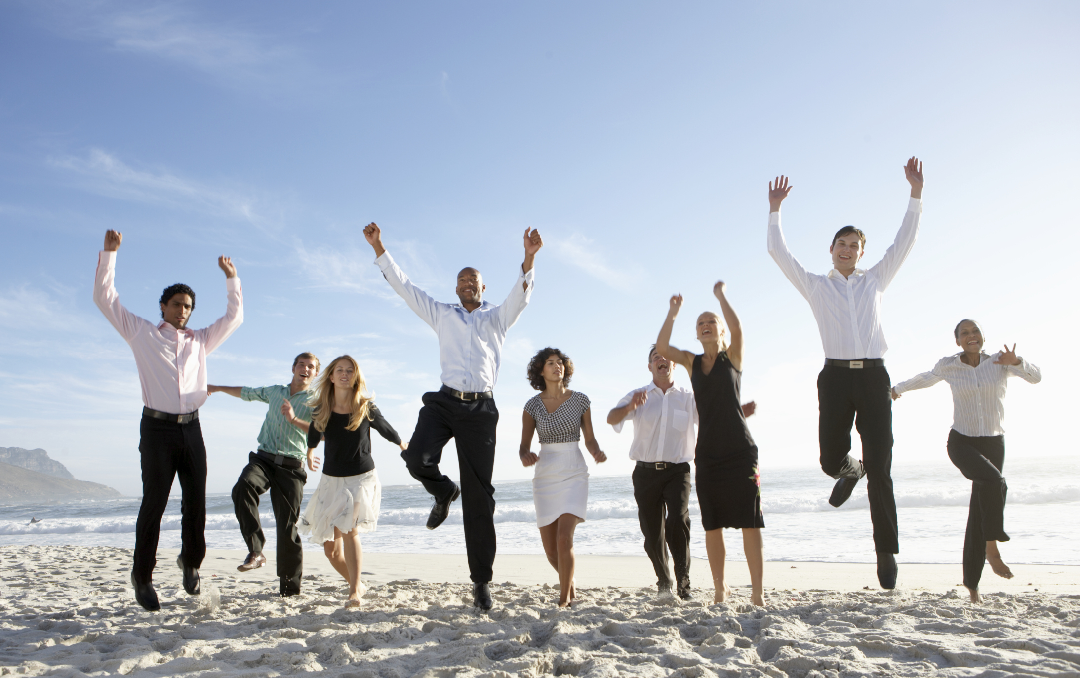 Men and women in professional attire jumping for joy on a beach.