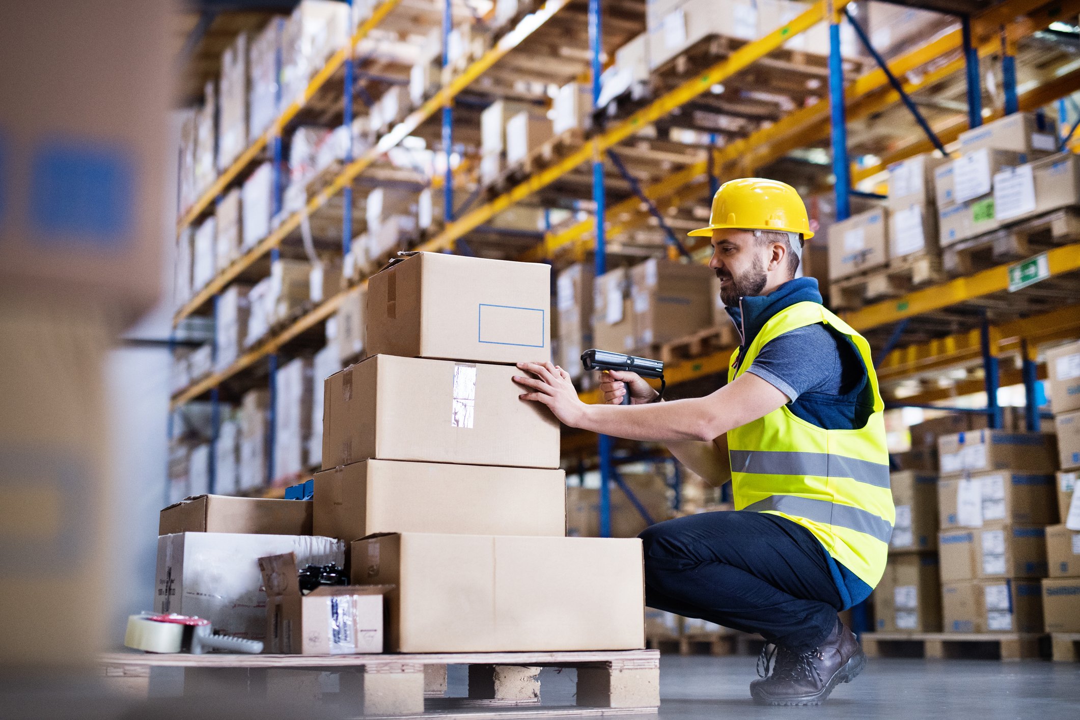 A man wearing a yellow hard hat while using a barcode reader and scanning packages in a warehouse.