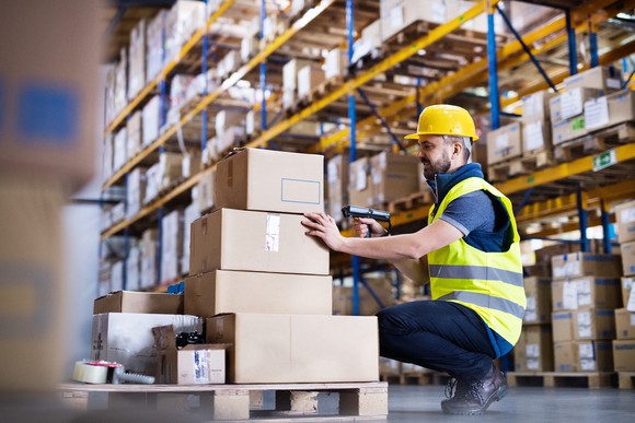 A man wearing a yellow hard hat while using a barcode reader and scanning packages in a warehouse.