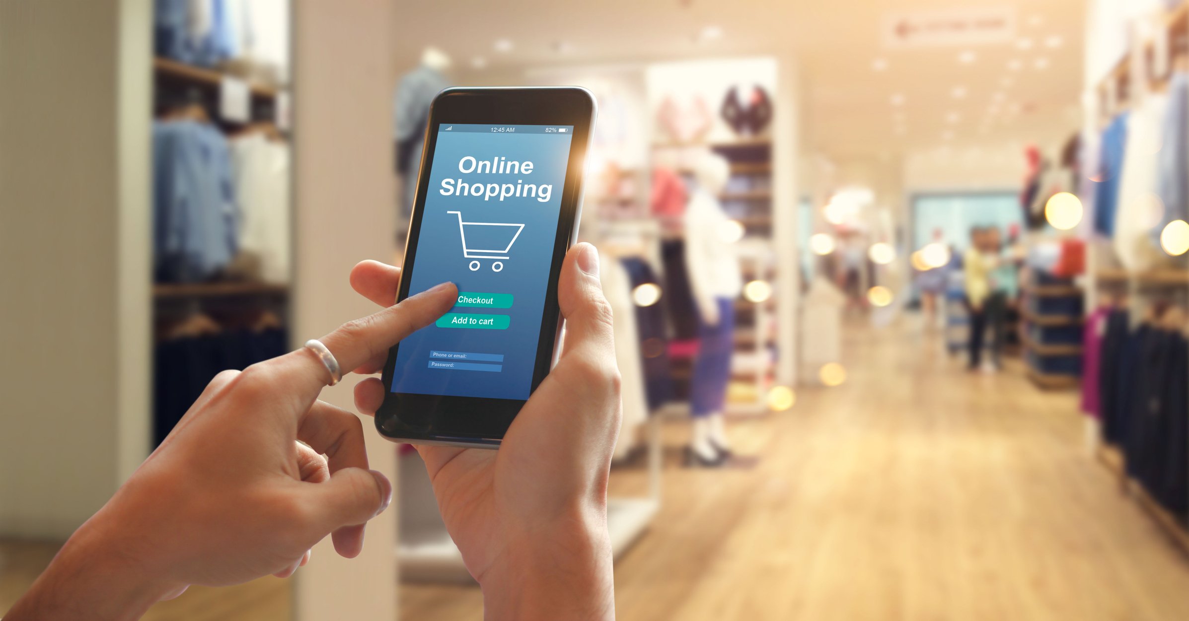 A woman's hands holding a smartphone with the words "online shopping" appearing on the screen and the interior of a retail store in the background.