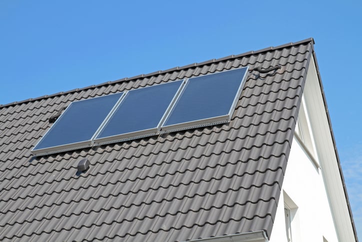 Three solar panels on a gray-brown roof of a white house with blue sky in background.