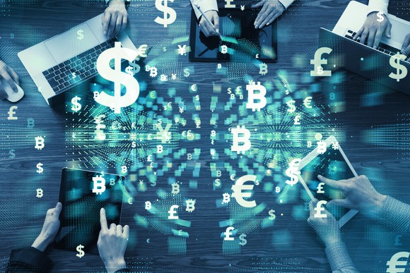 People sitting around a table with laptops and tablets, with digital symbols for currencies floating above table.