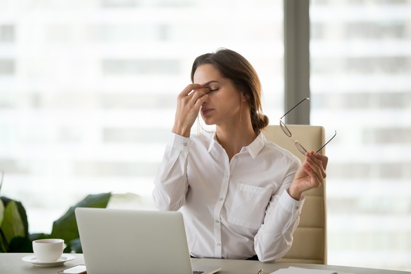 Woman in collared shirt at a laptop holding her face as if tired