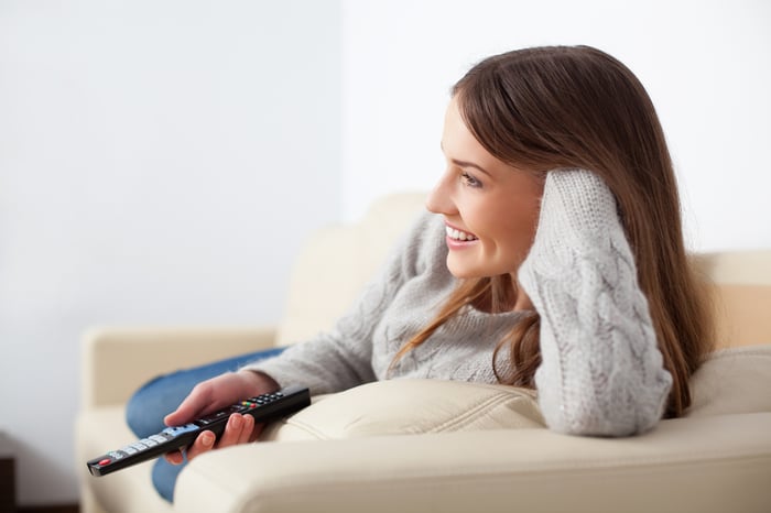 A smiling young woman wielding a TV remote on a beige couch.