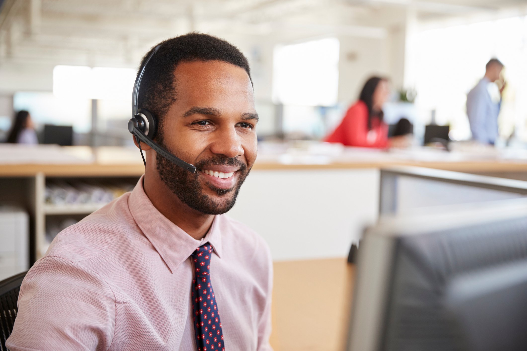 Smiling man wearing headset, and sitting in front of computer in an office.