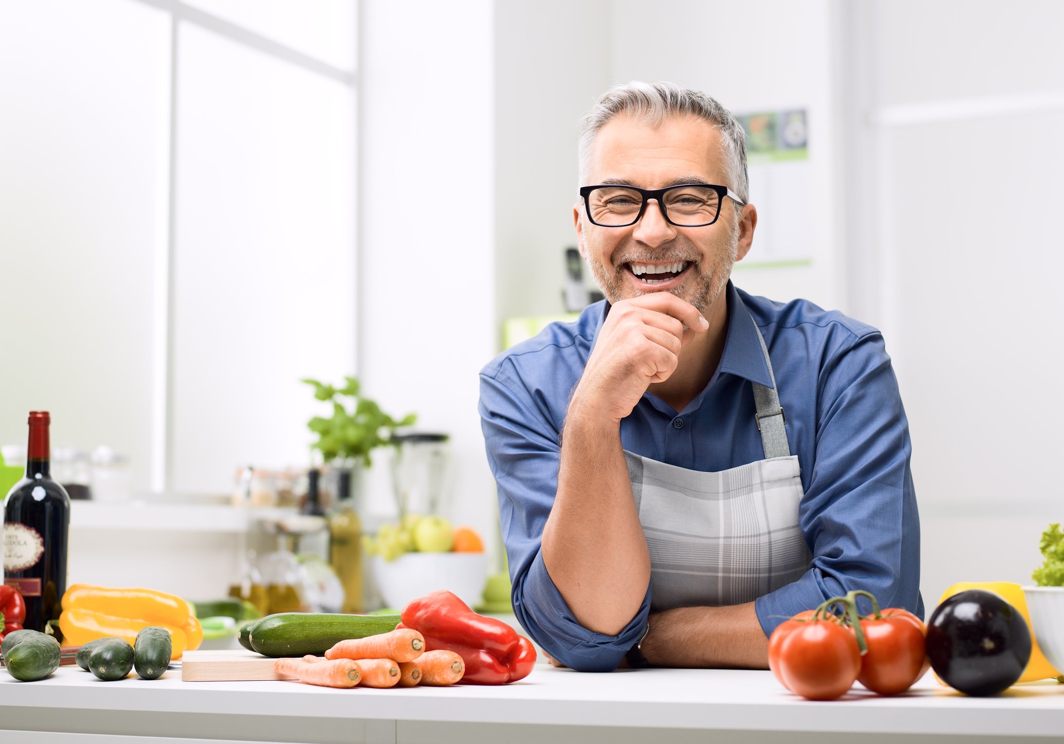 Older man in apron smiling, leaning against counter with various vegetables on it