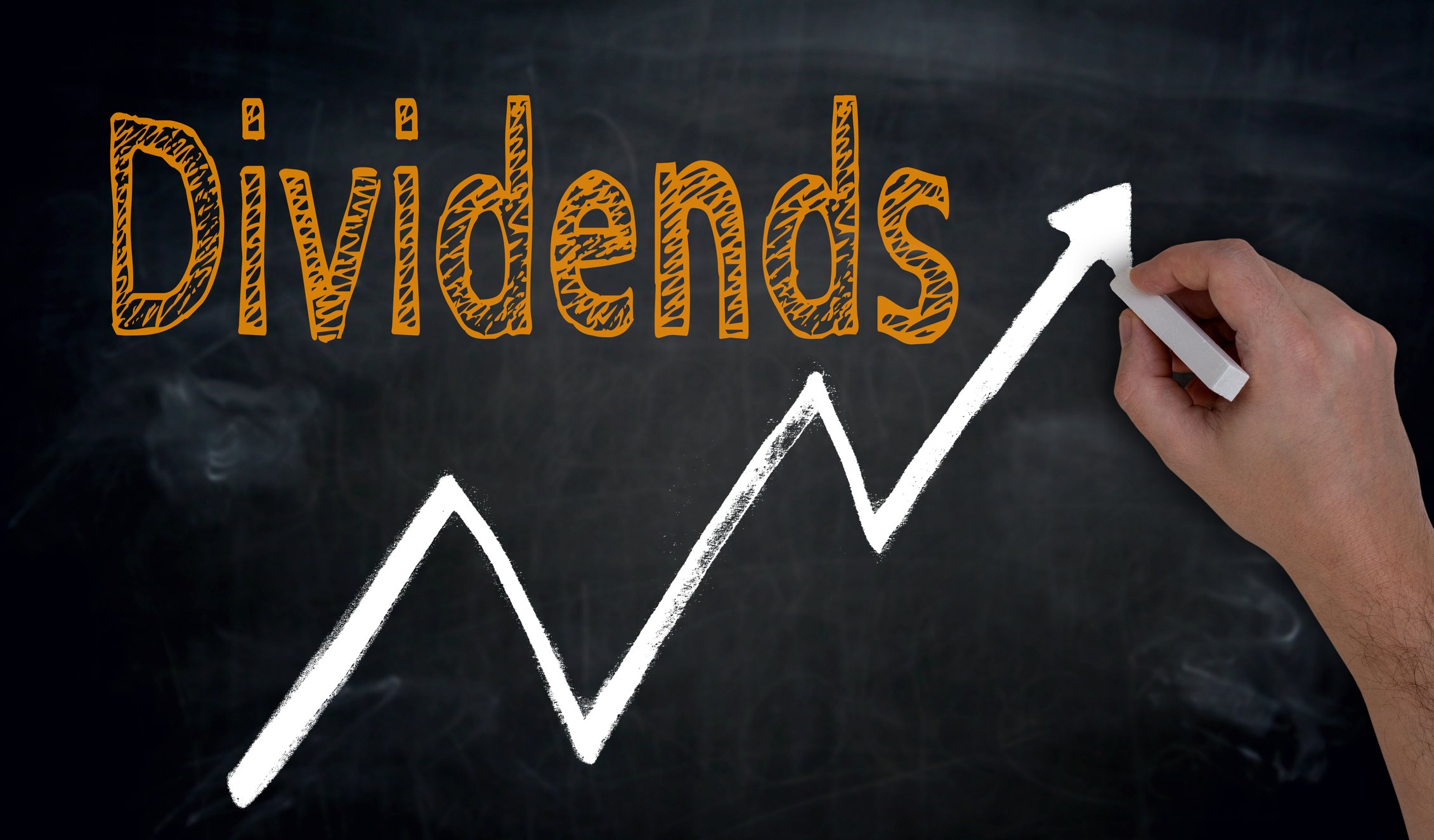 A hand draws an upward sloping line on a chalkboard with the word dividends in orange chalk above it. 
