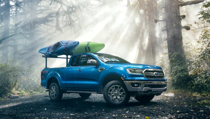 A blue 2019 Ford Ranger, a midsize pickup, is shown in a forest setting. There are two kayaks loaded on a rack in the Ranger's bed. 