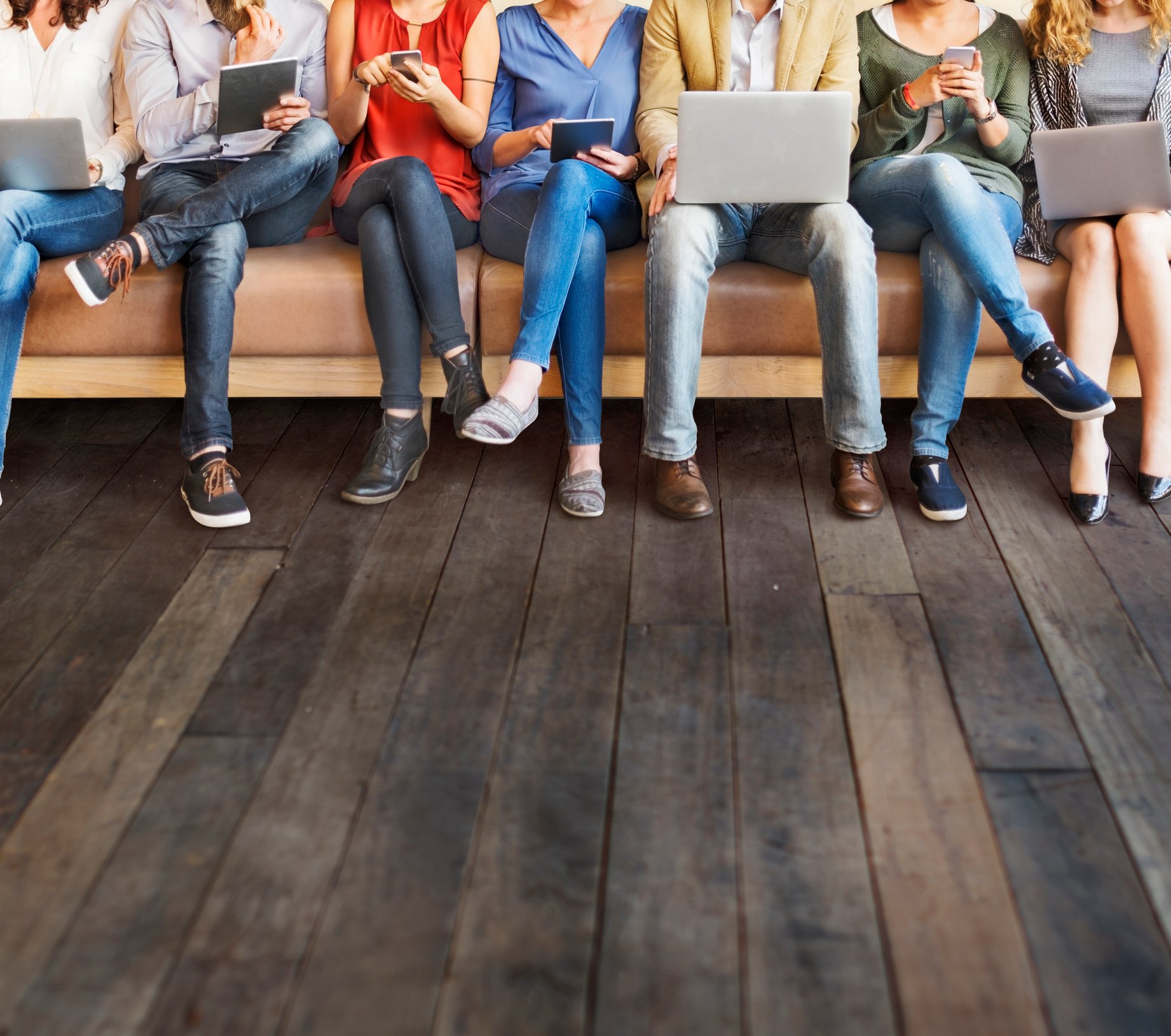 A group of people sitting on a bench, all holding laptops, tablets, or smartphones.