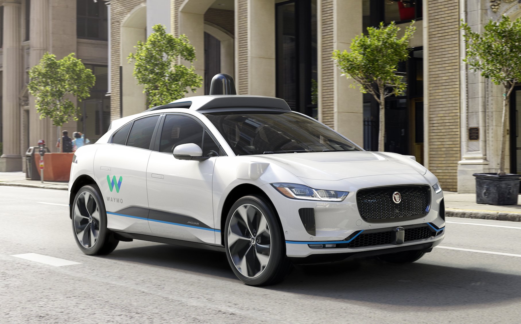 A white Jaguar I-Pace SUV, with self-driving sensor hardware and Waymo logos, on a city street. 
