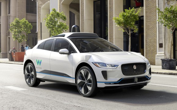 A white Jaguar I-Pace SUV, with self-driving sensor hardware and Waymo logos, on a city street. 