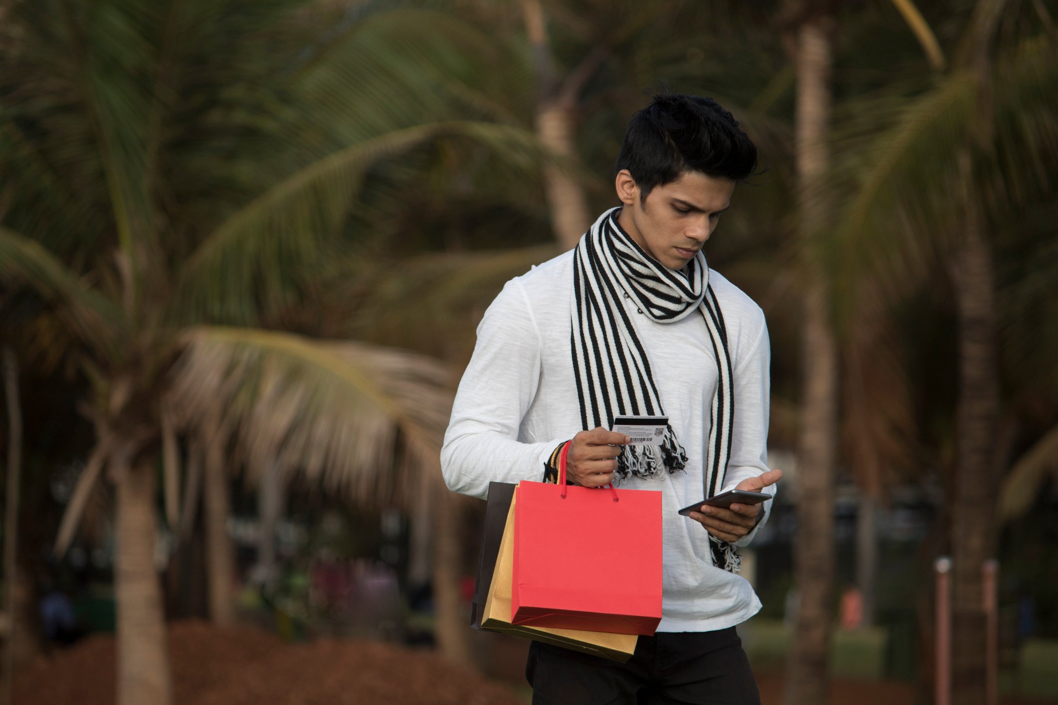 A young man with red and yellow bags in one hand looks down at his phone in the other.