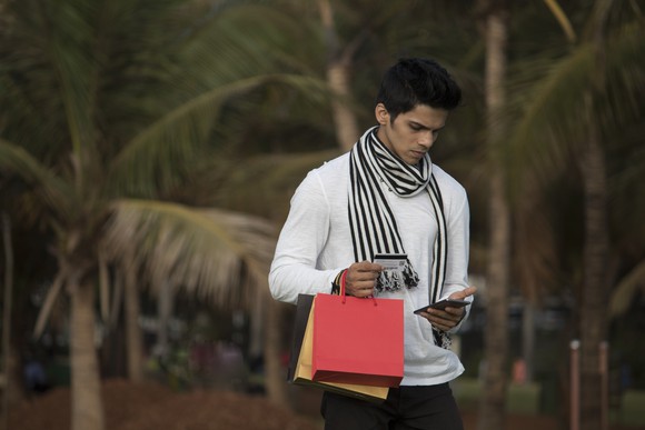 A young man with red and yellow bags in one hand looks down at his phone in the other.