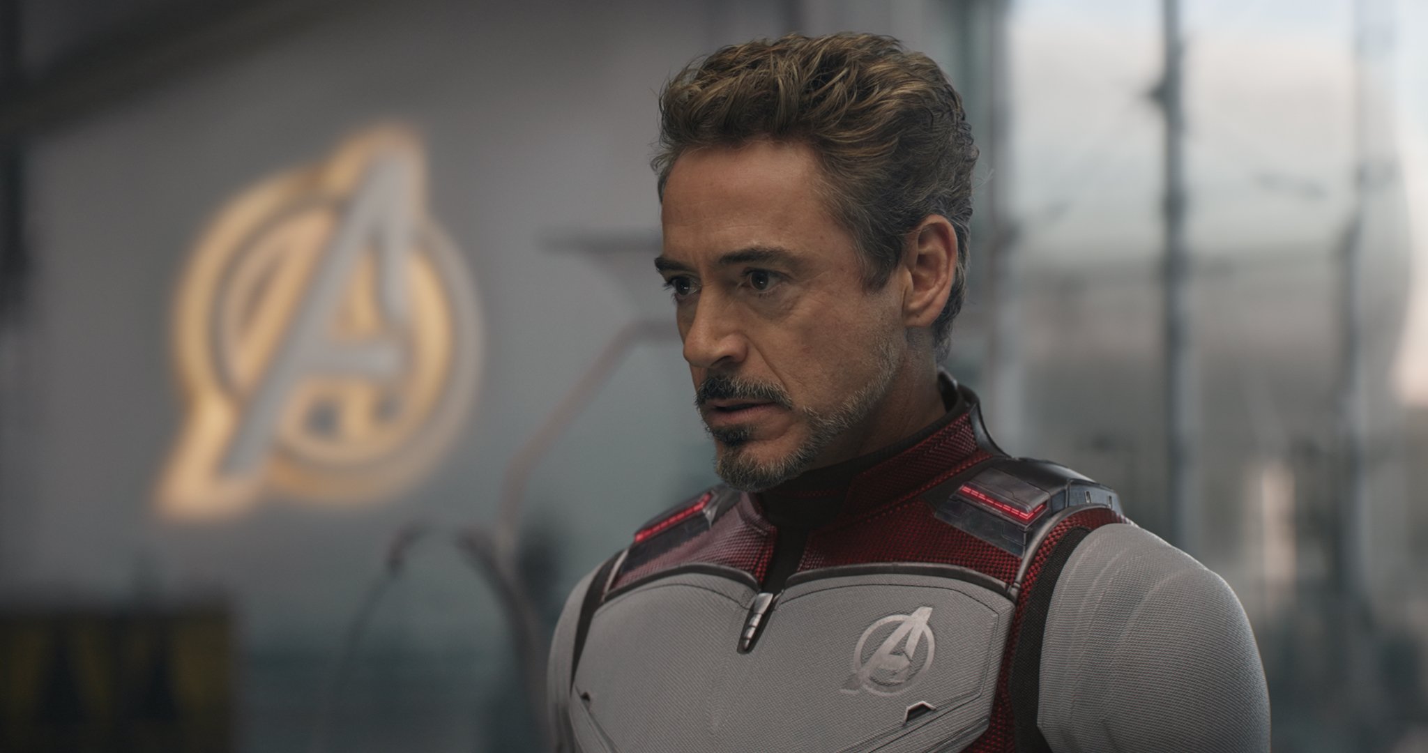 Iron Man stands near a lighted Avengers emblem, looking concerned. 
