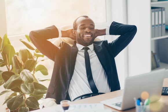 With his hands behind his head while sitting at a desk, a smiling man in a suit looks at his computer.