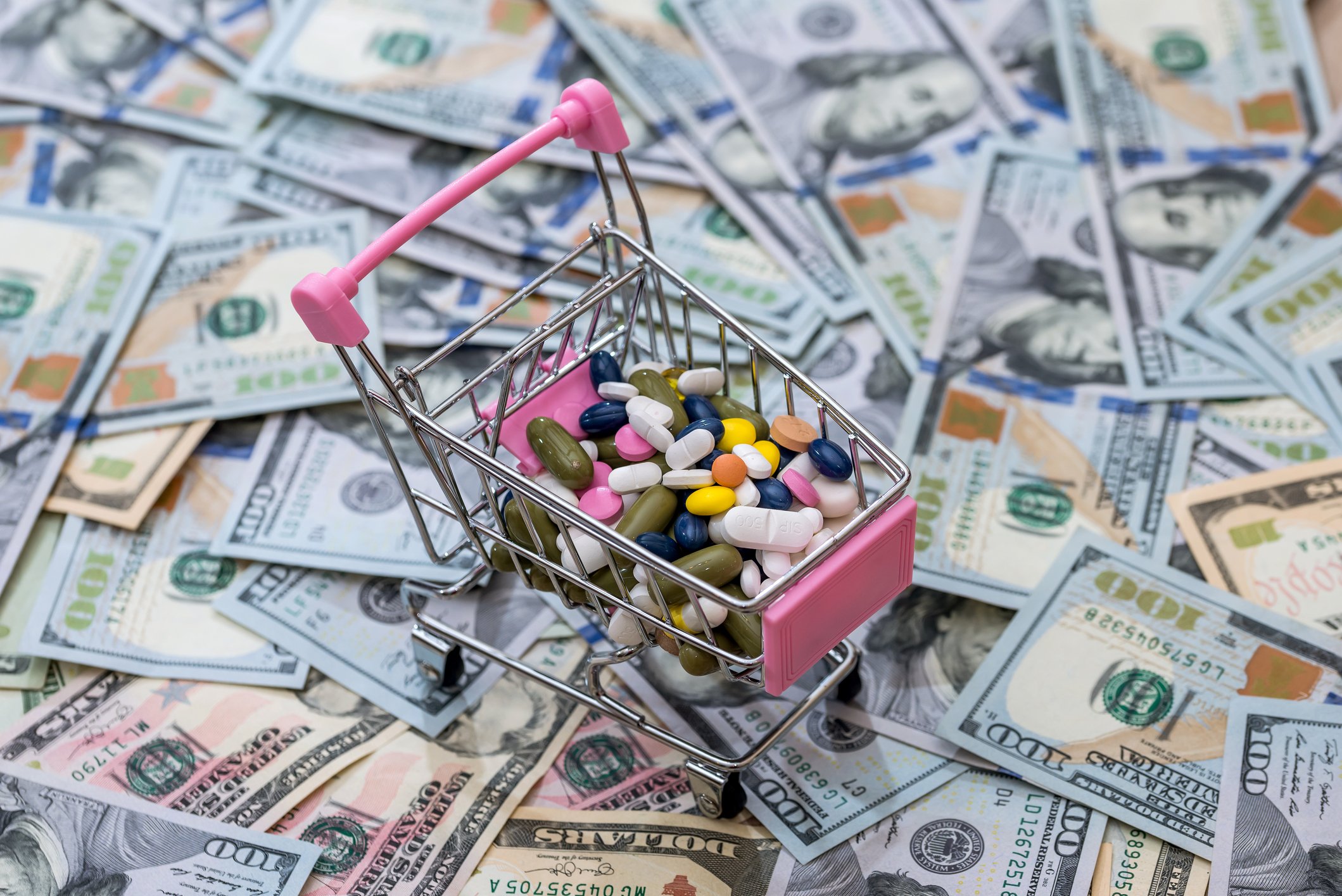 A miniature shopping cart loaded with various pills sitting on top of a stack of hundred-dollar bills.