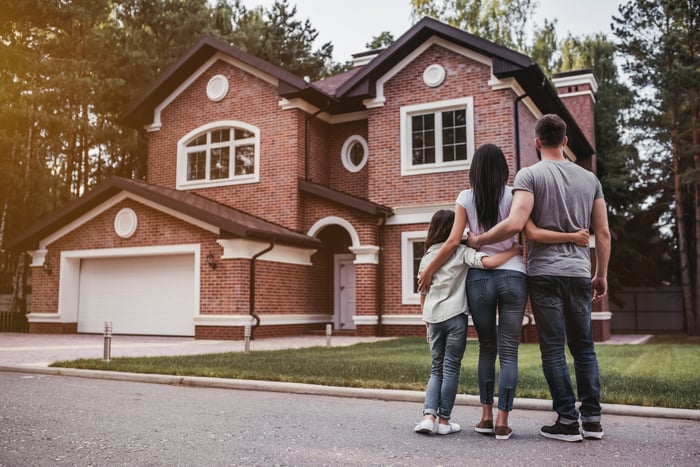 A man, woman, and child stand in the street with their arms around each other and look at a house.