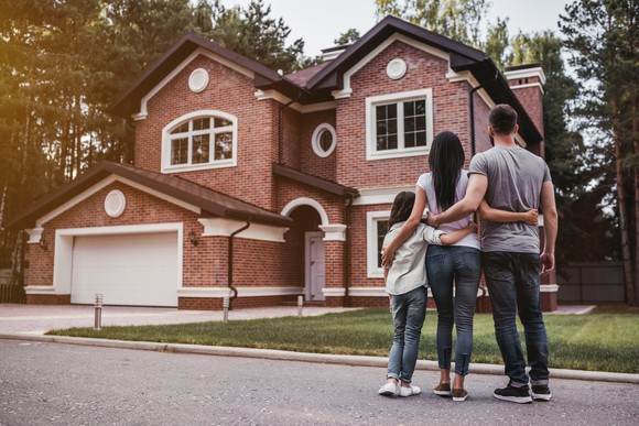 A man, woman, and child stand in the street with their arms around each other and look at a house.
