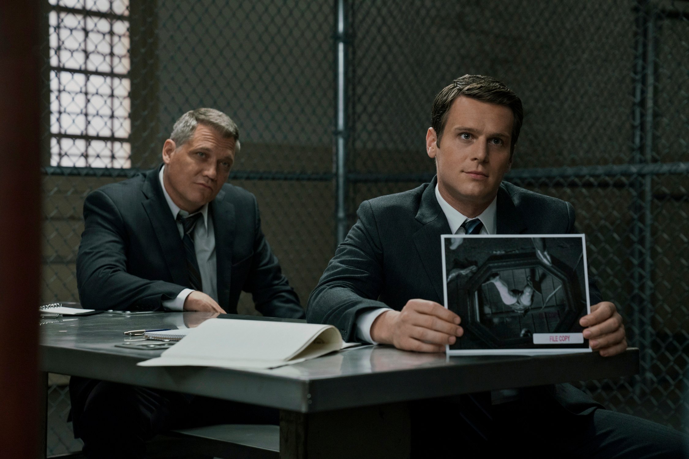 Two men in suits in a prison interrogation room showing a photo to an unseen inmate. 