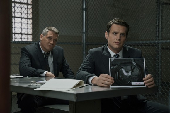 Two men in suits in a prison interrogation room showing a photo to an unseen inmate. 