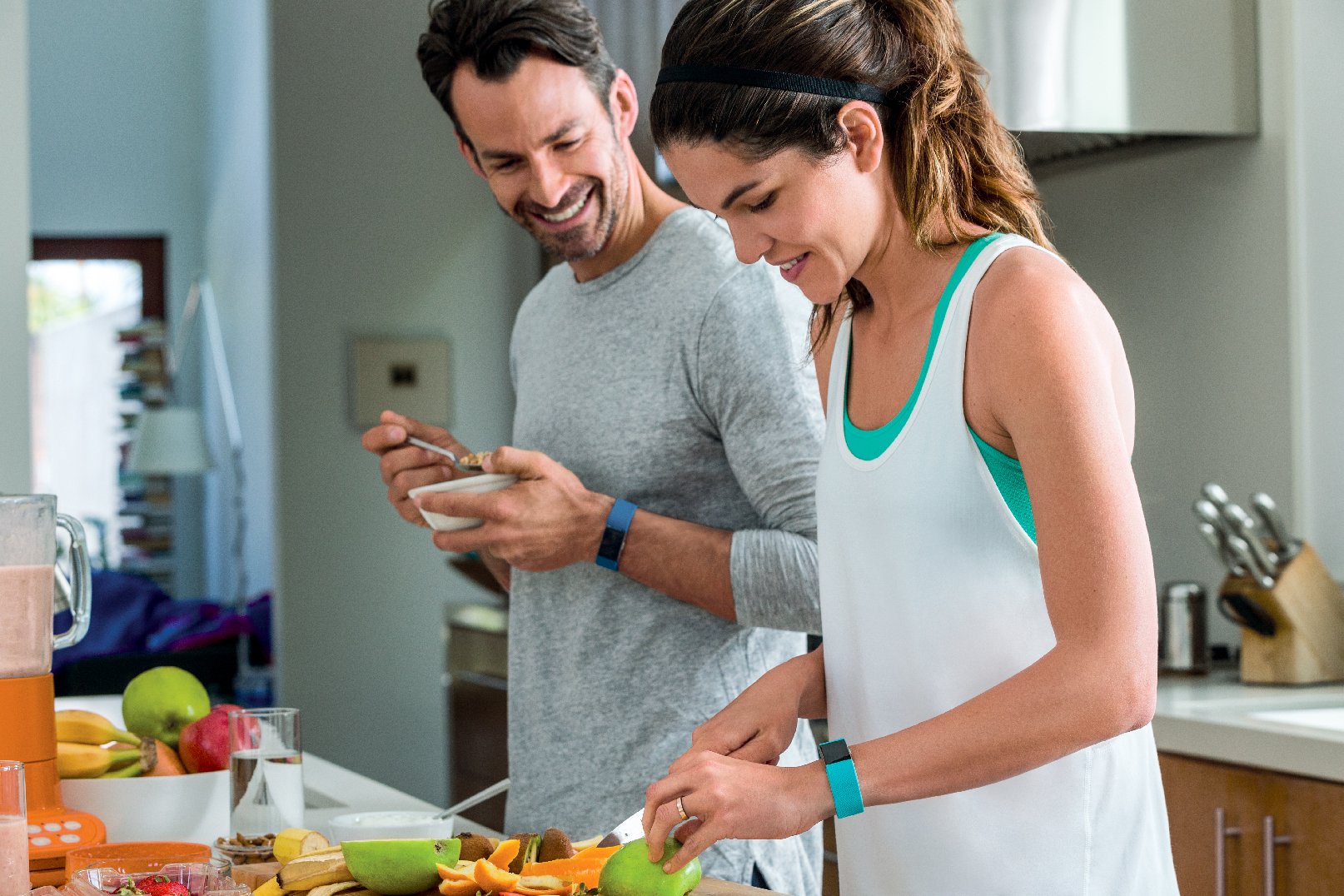 A couple wearing Fitbit Charge 2 trackers while cutting up fruit in a kitchen.