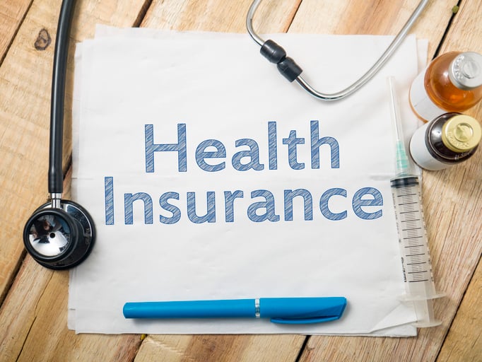 Piece of paper, with the words health insurance written on it, lying on wood surface with medical items (stethoscope, medicine) surrounding it.