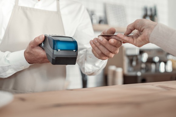 A hand hands a credit card to a cafe barista holding an electric point-of-sale device in his hand. 