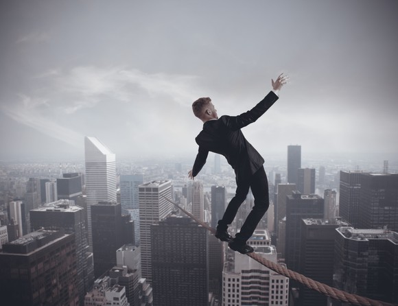 Businessman balancing precariously on a wire streatched high above a bustling cityscape.