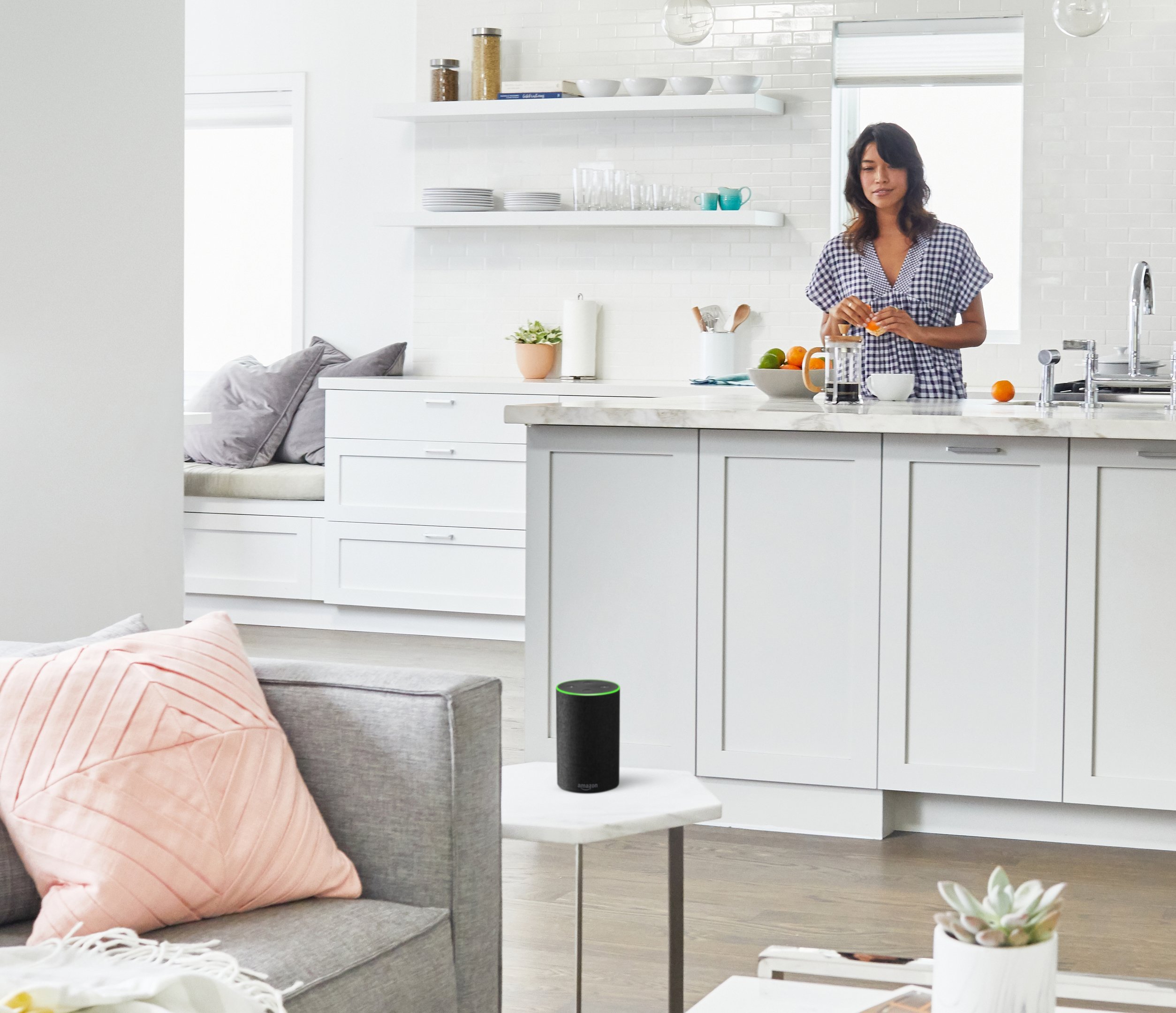 A woman peeling an orange at the kitchen counter, looking at an Echo smart speaker on a small table in the adjacent living room.