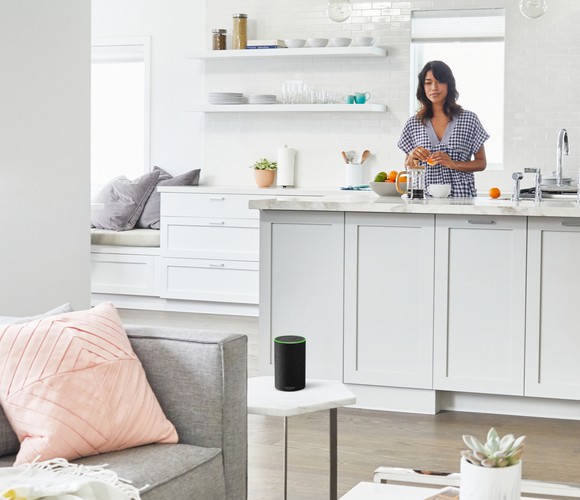 A woman peeling an orange at the kitchen counter, looking at an Echo smart speaker on a small table in the adjacent living room.