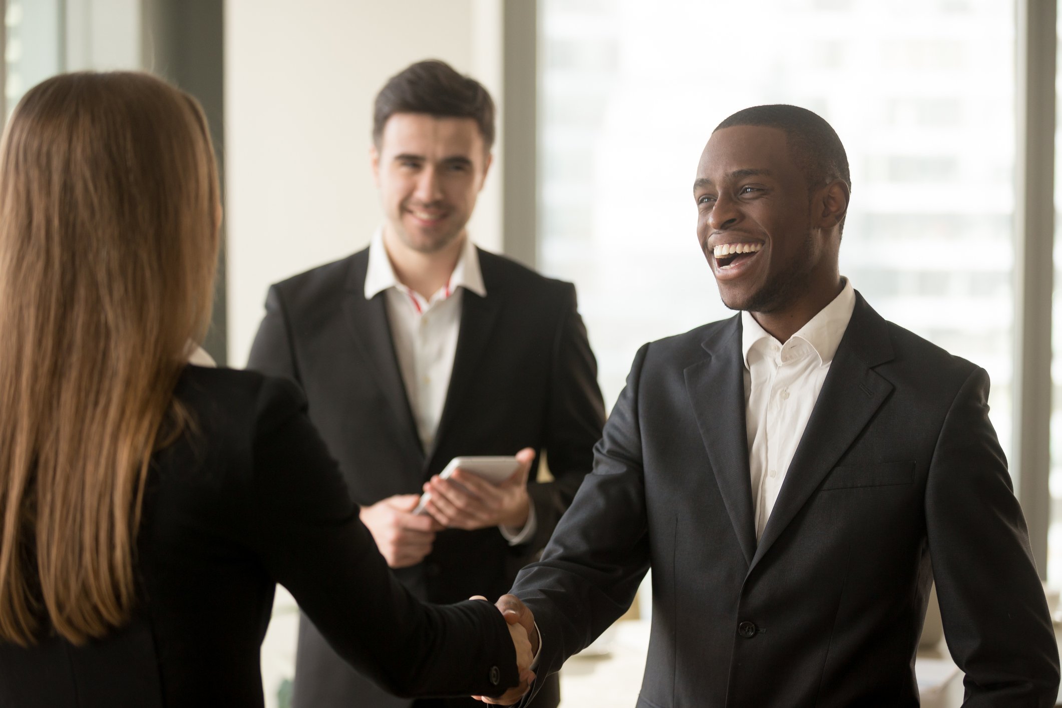 A smiling man in a suit shaking hands with a woman in a suit, while another man in a suit is looking on.