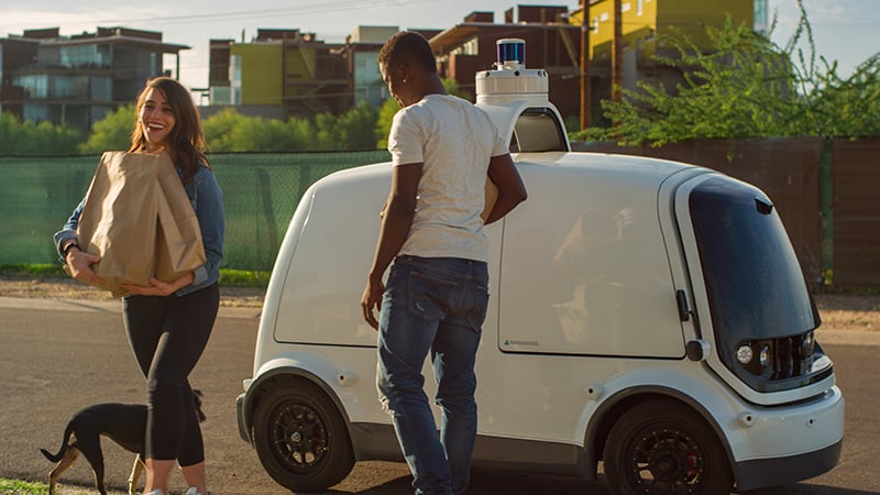 Two people retrieving grocery bags from a Nuro self-driving vehicle, a white box-shaped car with doors and a sensor array on top.