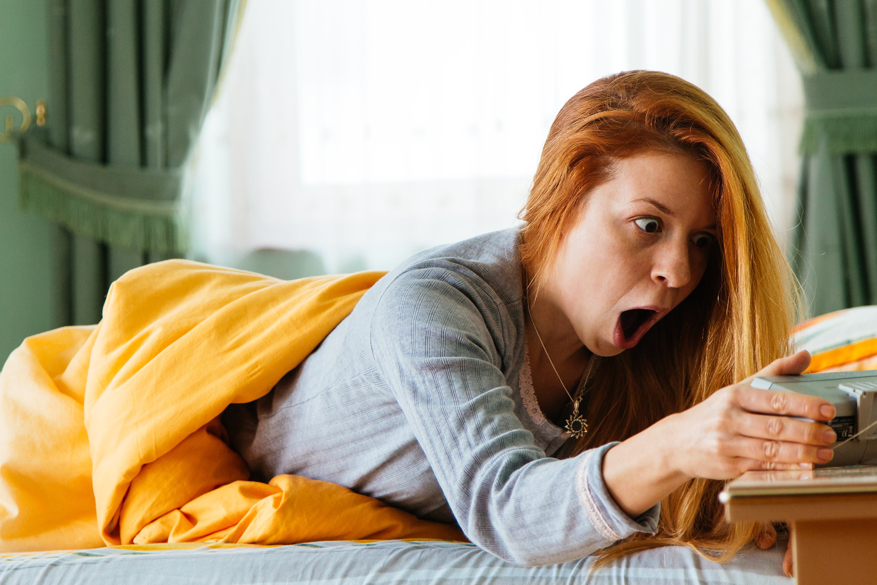 We see a woman in bed grasping her alarm clock with her mouth wide open in shock and alarm.