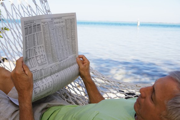 A man resting in a hammock near the ocean reading a newspaper.