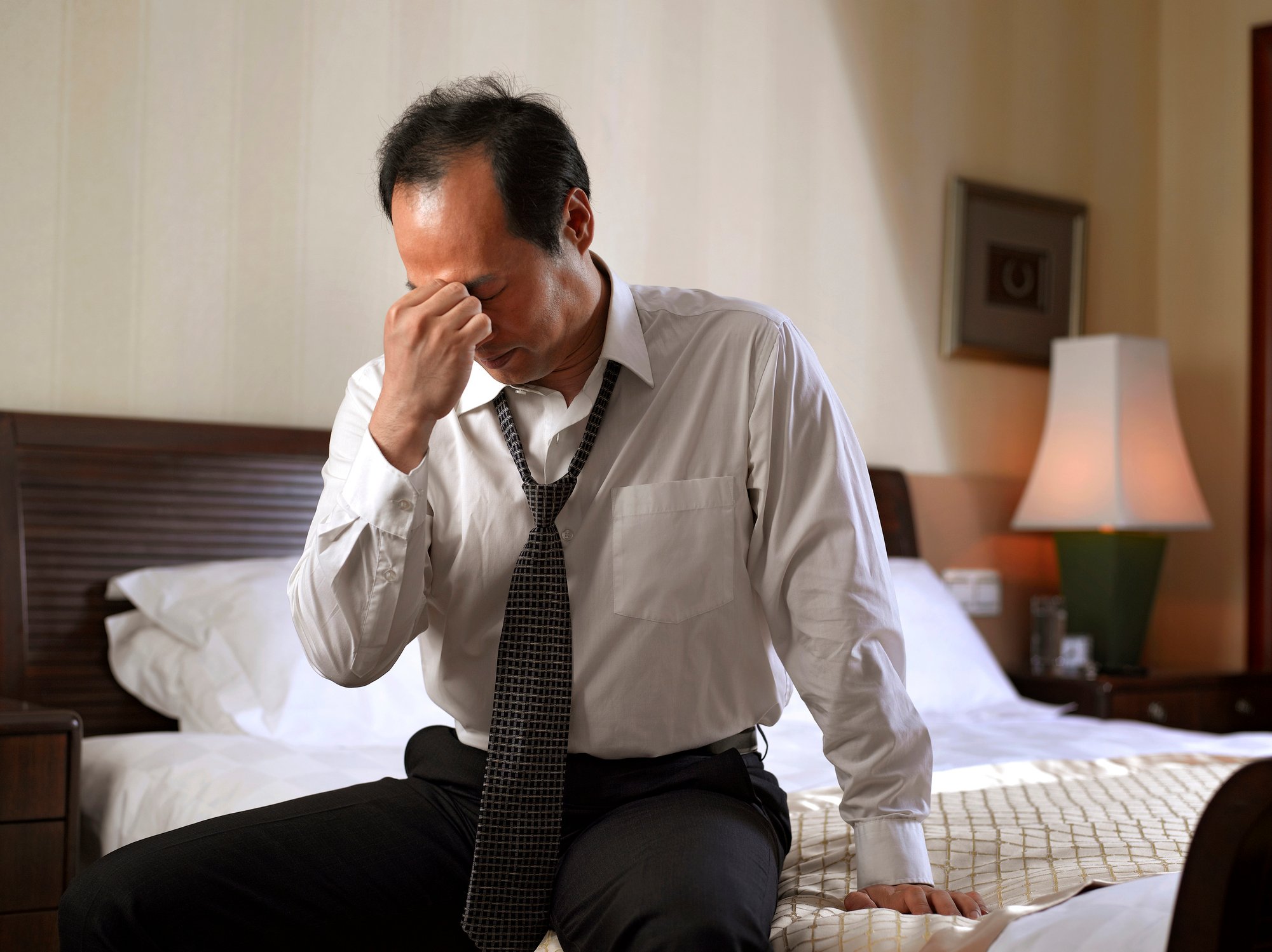 Man dressed in professional attire, sitting on the edge of a bed and pinching his face