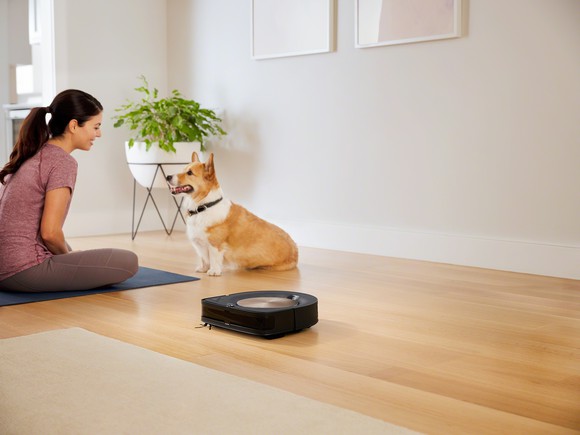 A woman, who's sitting on a yoga mat, looking at her dog while the iRobot Roomba S9 robotic vacuum cleans the wooden floor in the foreground.