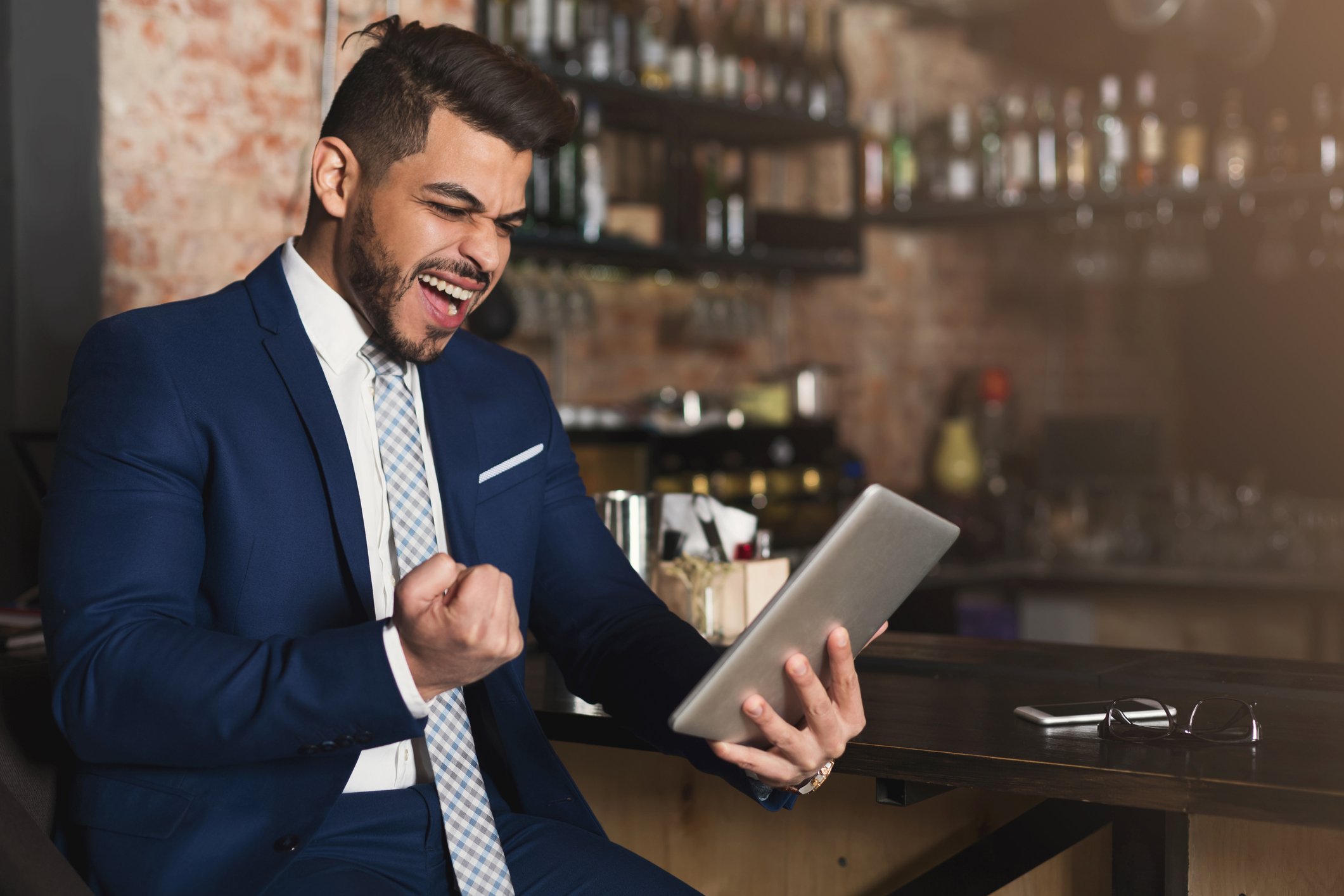 A man pumps his fist in joy while looking at something on his tablet in a bar. 