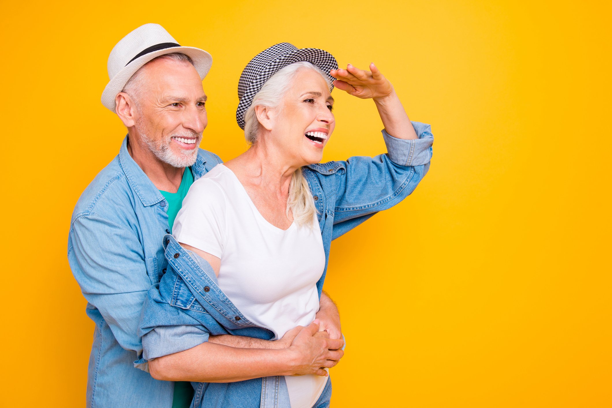A senior couple wear cool thin bill hats and denim shirts as the woman looks into the distance against an orange background.