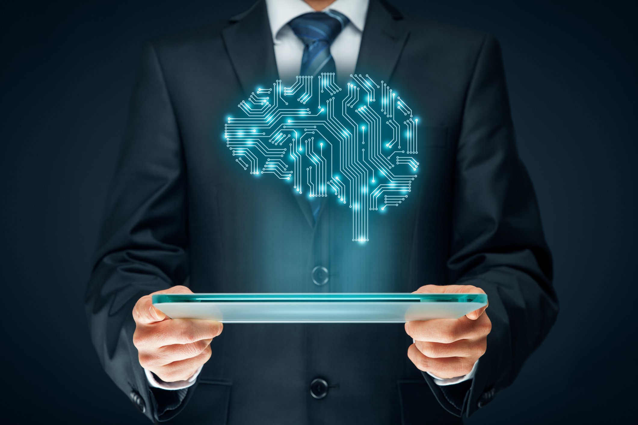 A man in a suit holding a tablet with an illustrated brain made of electrical connections hovering above it, illustrating artificial intelligence.