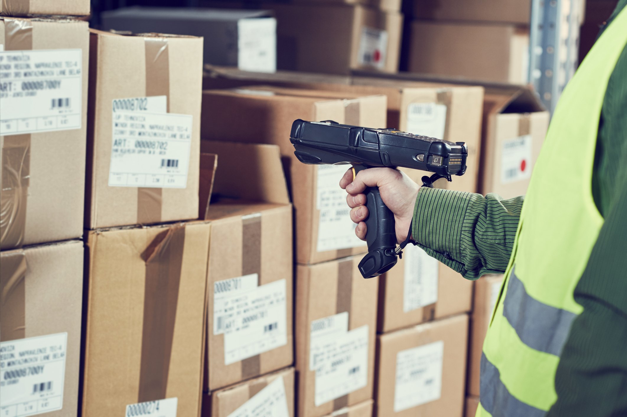 A warehouse worker using a handheld barcode scanner to read codes on a large box that's surrounded by many other boxes.