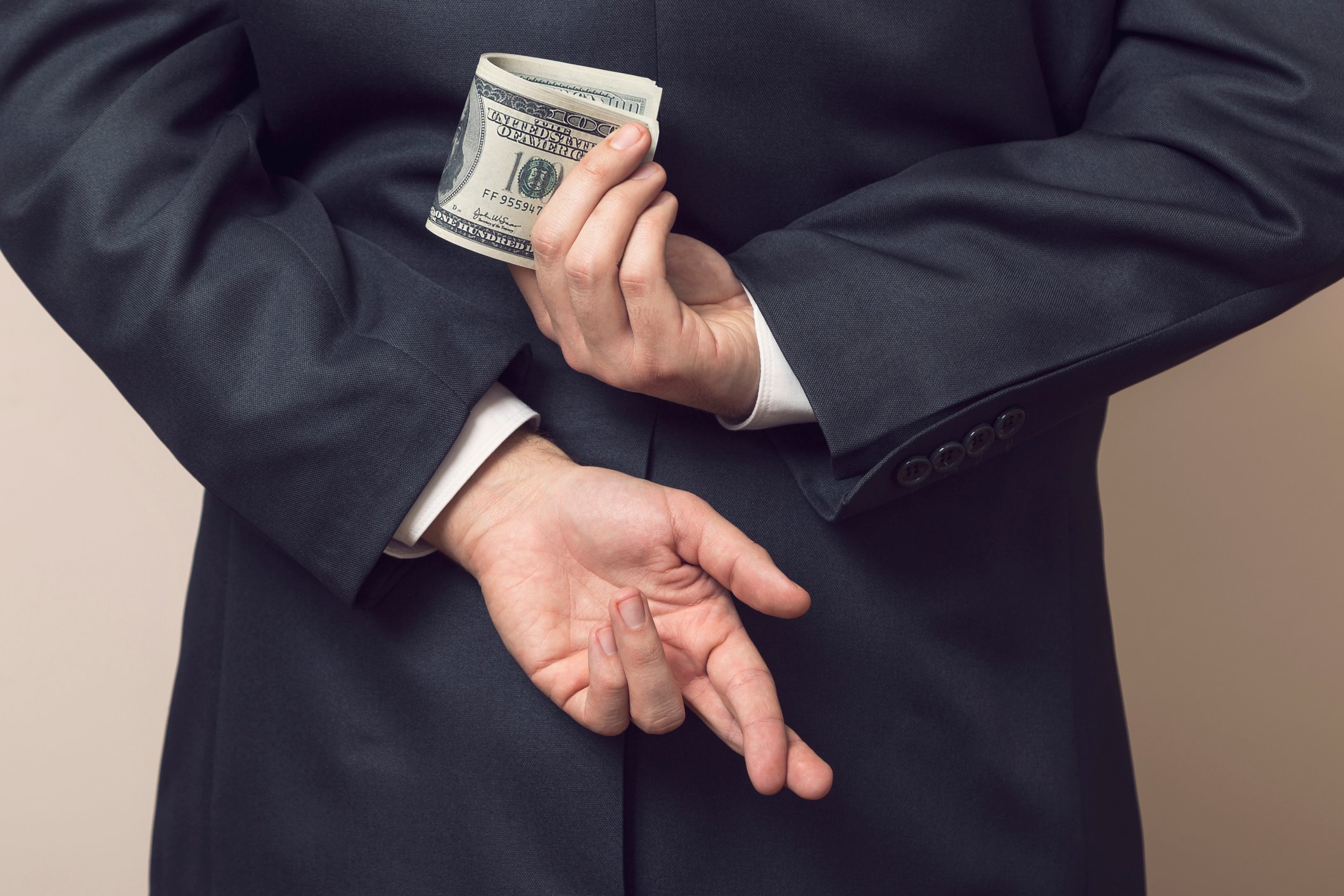 A businessman crosses his fingers behind his back while holding a wad of hundred dollar bills.