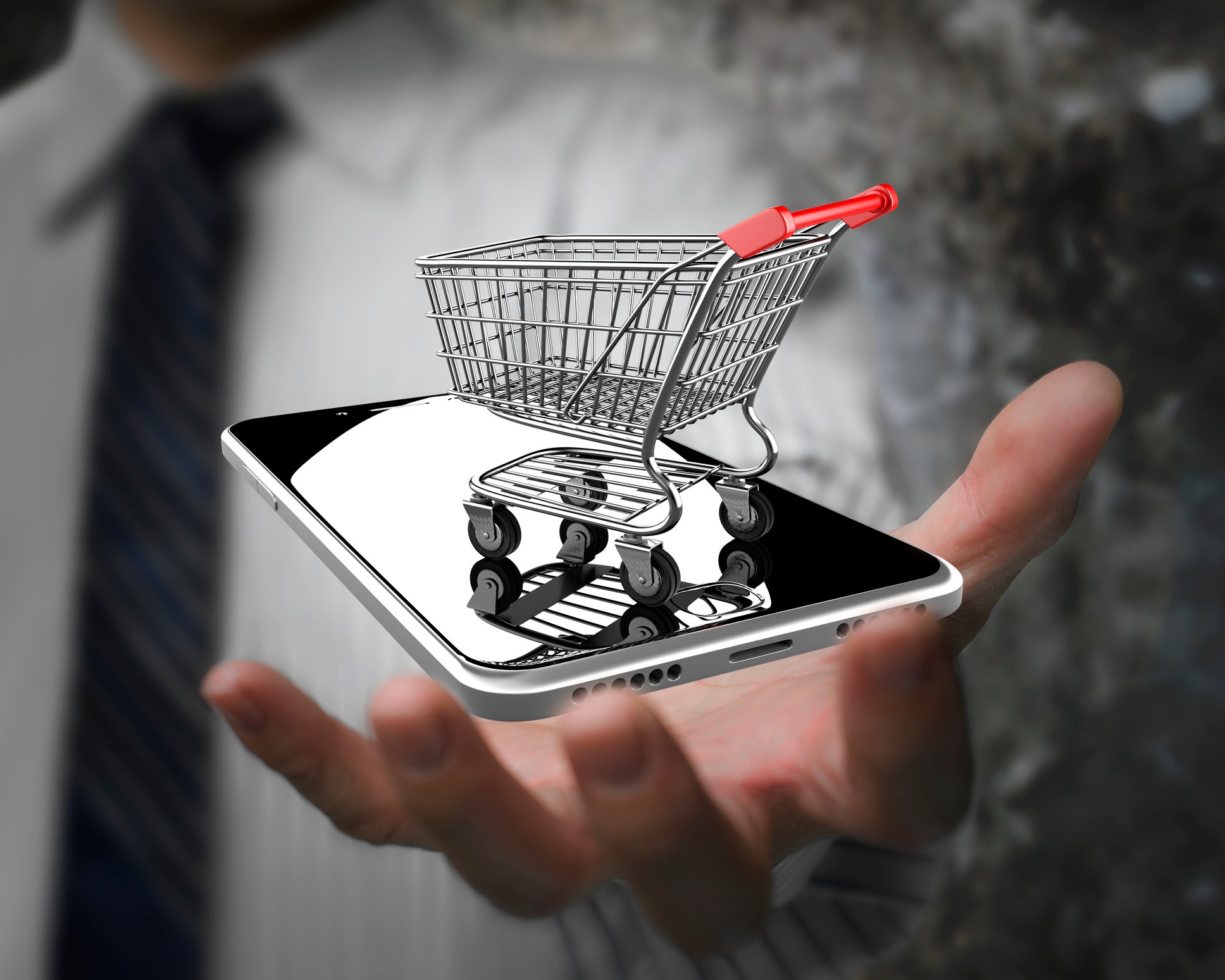 A man holding a mobile phone that has a miniature shopping cart on top of it.