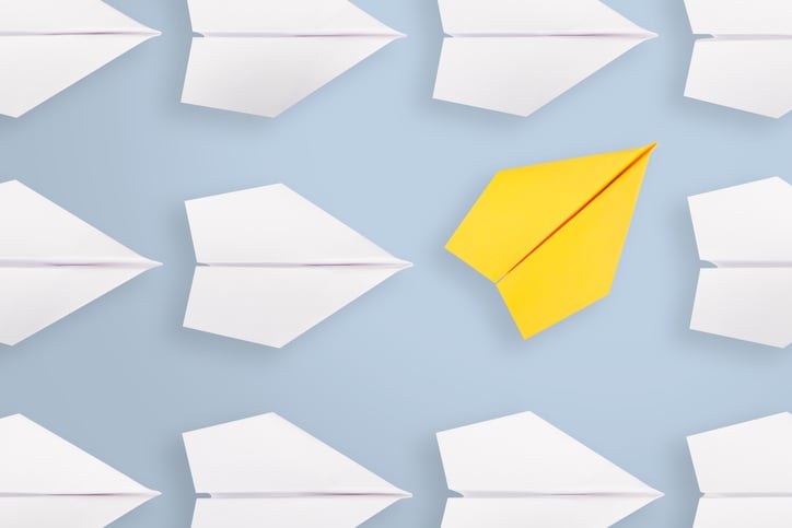 A yellow paper airplane moving in a different direction than many white paper airplanes nearby.