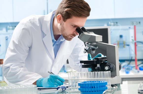 A man in lab coat and gloves looking through a microscope.