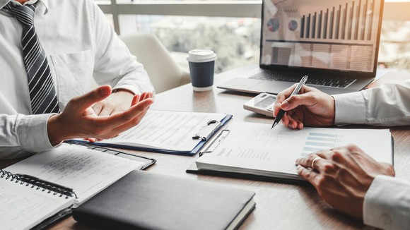 With a financial chart displayed on a computer behind them, two businessmen work at a desk.
