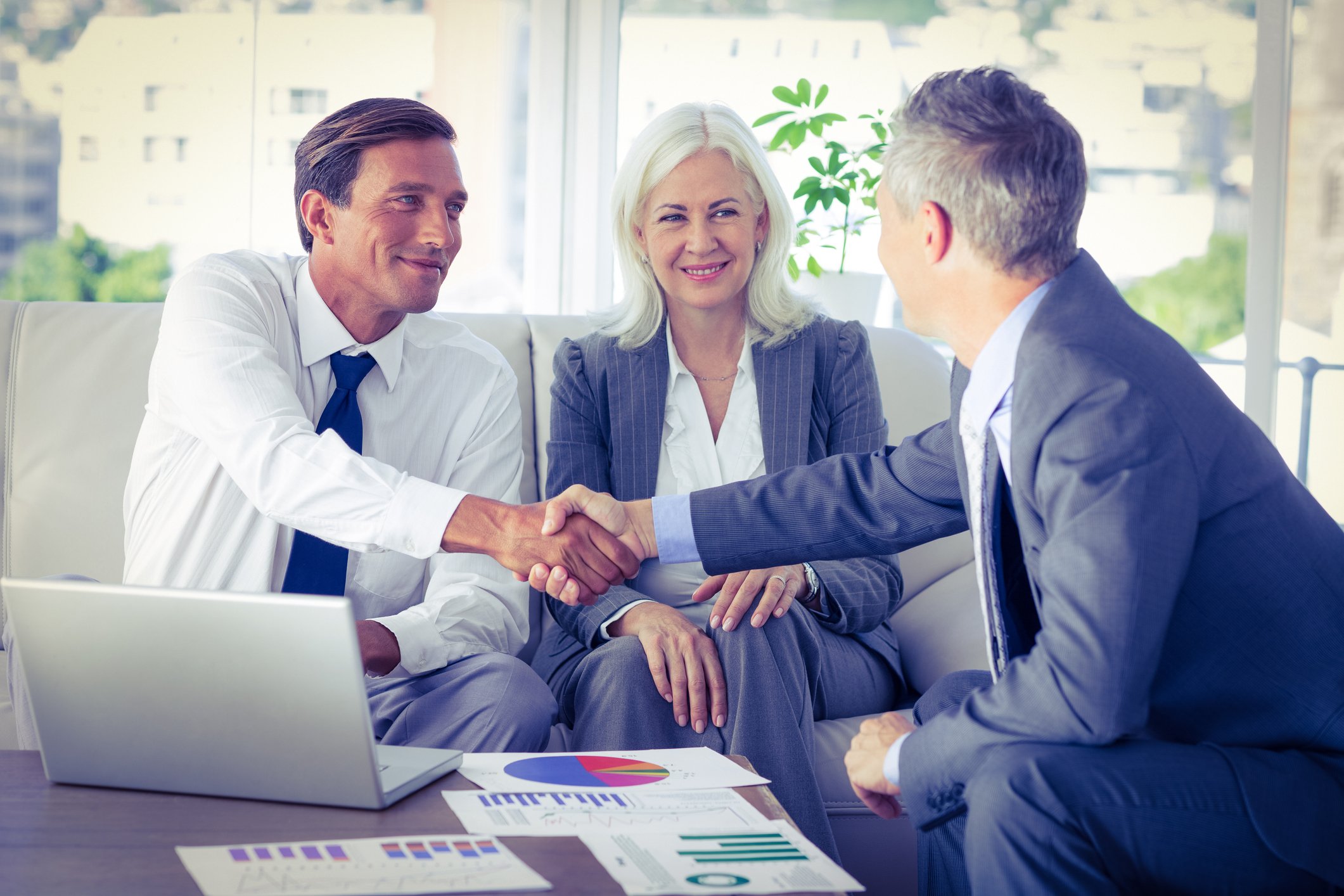 Three businesspeople on a couch looking at a laptop and some charts; the two men are shaking hands while the woman looks on.