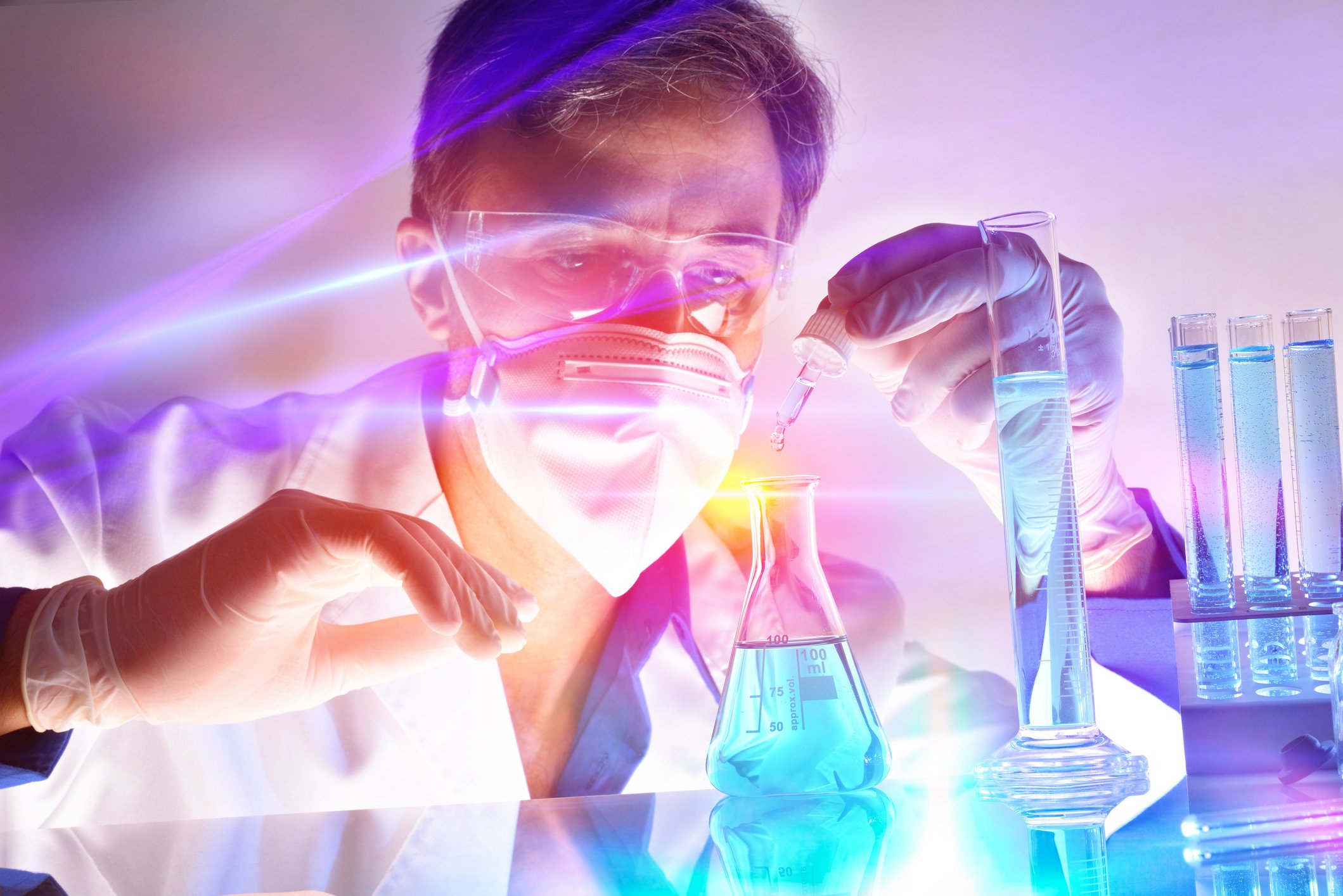 Man in lab coat, goggles, and face mask working with a beaker and test tubes.