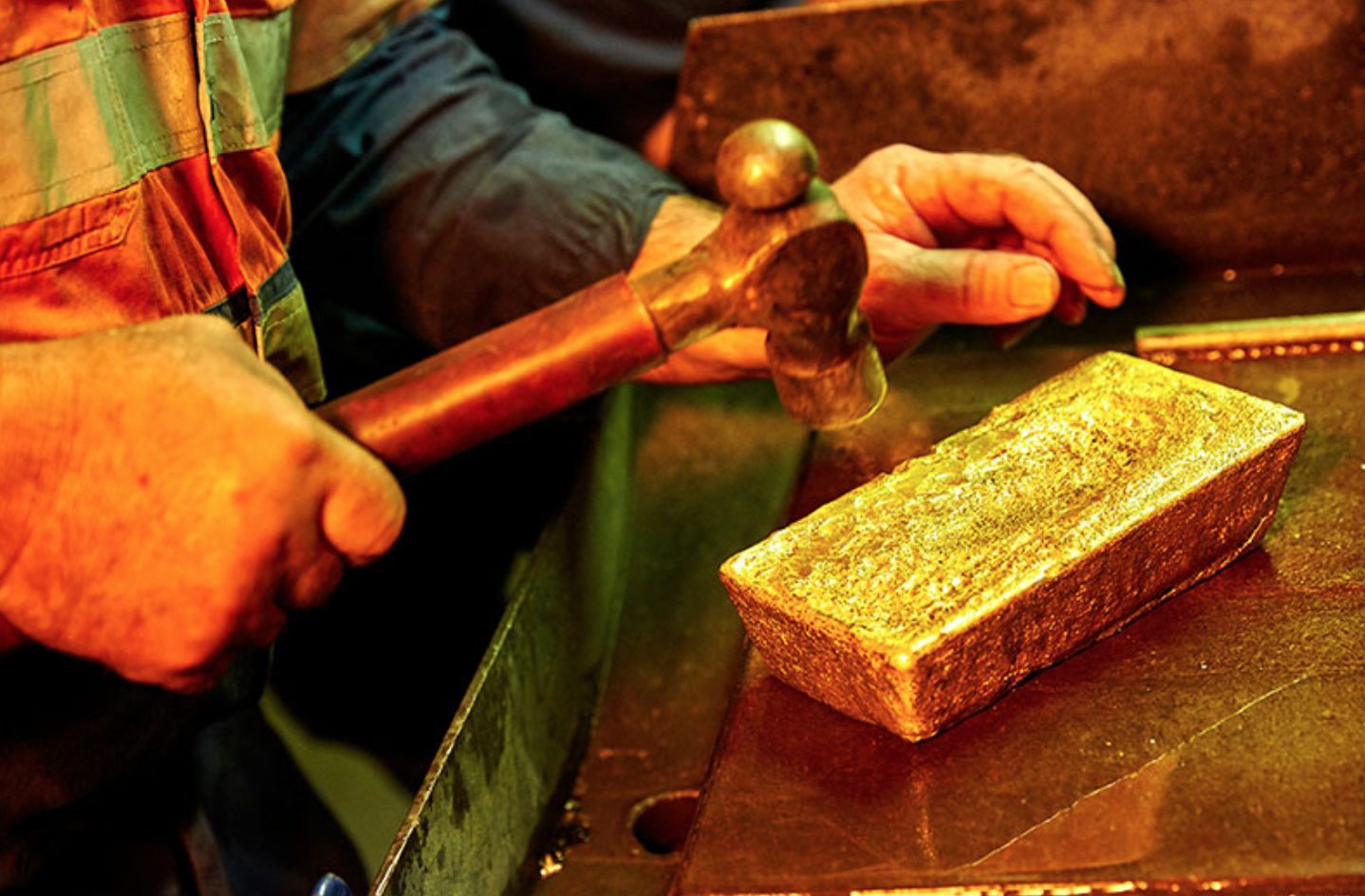 A worker holds a hammer over a bar of gold.